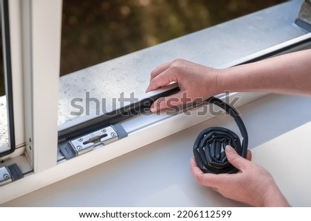 Preparing for the cold season. A woman sticks a dark rubber sealing tape on a window indoors.
