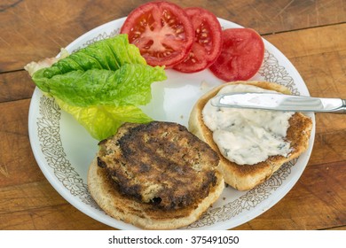 Preparing Crab Cake On Toasted Bun With Sliced Tomatoes And Lettuce.  Tartar Sauce Being Spread Over Bun. Overhead View.