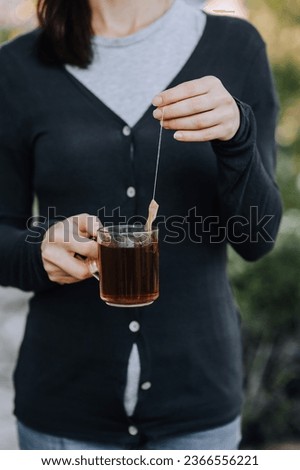 Preparing a delicious drink, hot tea in a bag in a glass cup in the hands of a woman outdoors. Food photography, close-up portrait.