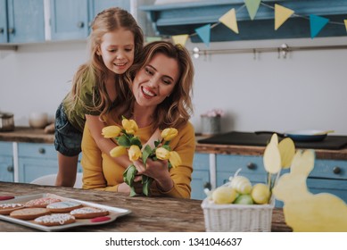 Preparing for Easter holiday. Waist up portrait of happy female child embracing her beautiful smiling mom with bunch of yellow tulips while being on festive kitchen Stock Photo