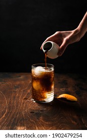 Preparing espresso tonic with orange juice. A hand pouring espresso from a small white can into a highball glass filled with ice spheres and tonic. 