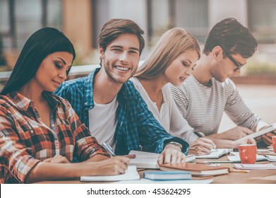 Preparing to exams together. Joyful young man smiling and looking at camera while sitting with his friends at the wooden desk outdoors - Shutterstock ID 453624295