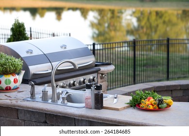 Preparing a healthy summer meal in an outdoor kitchen with gas barbecue and sink on a brick patio overlooking a tranquil lake with tree reflections