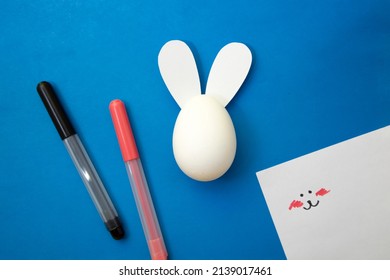 Preparing For The Holiday Of Happy Easter. Coloring Book For Children. White Easter Egg With Bunny Ears Glued On. Markers On A Blue Background. White Sheet With Coloring Example.