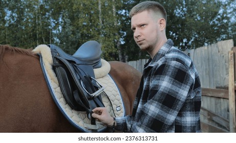 Preparing for a horseback ride. Saddling a horse by a young man. The rider saddles the horse and prepares to ride on the ranch. Horse racing, a male jockey prepares a horse in a paddock. - Powered by Shutterstock