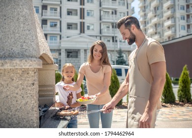 Preparing Meat. Dad Grilling Meat While His Daughter And Wife Standing Next To Him