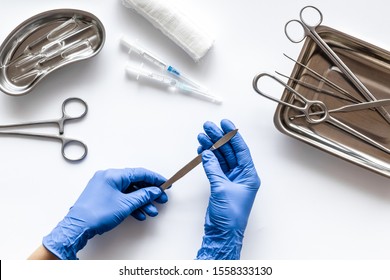 Preparing For Plastic Surgery. Doctor's Hands Takes Scalpel On White Background With Surgical Tools Top View