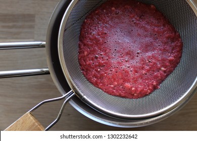 Preparing Pomegranate Puree By Separating Juice From Pulp And Seeds With The Use Of A Fine Mesh Strainer. Homemade Grenadine Preparation. Top View Closeup Of Thick Red-pink Mash And Wood Background