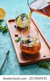 Preparing refreshing drink, iced tea with lemon slices, mint and blueberries in glasses on turquoise concrete background. Soft drinks. Summer drinks