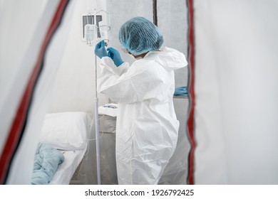 Preparing. Side View Of The Young Multiracial Woman Doctor Or Nurse Dressed In Protective Gown, Putting The Dropper In Hospital Room. Coronavirus Pandemic Concept. Stock Photo