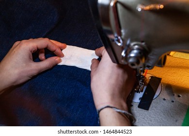 Preparing To Sow White Patch On Jeans. A Closeup View On The Hands Of A Young Fashion Designer Placing White Label On Blue Denim Garment Beneath Industrial Sewing Machine.