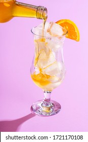 Preparing A Summer Cold Drink By Pouring Orange Syrup Over Ice Cubes In A Glass, On A Purple Background. Making Orange Lemonade. Tropical Mocktail.