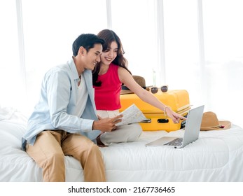 Preparing Travel And Vacation Planning Concepts. Young Pretty Asian Woman And Man In Denim Shirt Looking At The Laptop And Map On The Bed With Suitcase In Bedroom. Trip Planning On Summer Holiday.