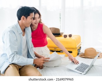 Preparing Trip And Vacation Plan Concepts. Young Pretty Asian Woman And Man In Denim Shirt Looking At The Laptop And Map On The Bed With Suitcase In Bedroom. Travel Planning On Summer Holiday.