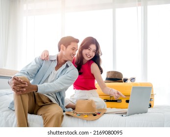 Preparing Trip And Vacation Plan Concepts. Young Pretty Asian Woman And Man In Denim Shirt Looking At The Laptop And Map On The Bed With Suitcase In Bedroom. Travel Planning On Summer Holiday.