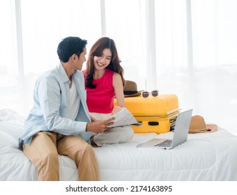 Preparing Trip And Vacation Plan Concepts. Young Pretty Asian Woman And Man In Denim Shirt Looking At The Laptop And Map On The Bed With Suitcase In Bedroom. Travel Planning On Summer Holiday.