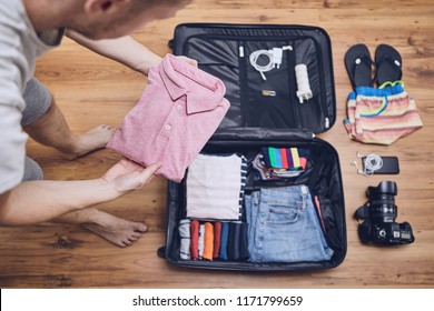 Preparing For Trip. Young Man Packing Clothing For Vacation, Flip Flop, Camera And Other Things On Hardwood Floor.