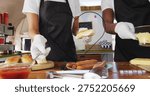 Preparing two servers assembling buns and dogs at counter, in white shirts and aprons, heart frame. Marketstall, streetfood, vendor, sausages, cheese, condiments, cuttingboard
