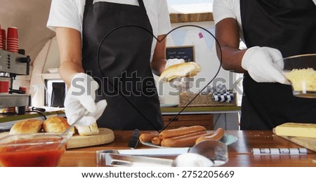 Preparing two servers assembling buns and dogs at counter, in white shirts and aprons, heart frame. Marketstall, streetfood, vendor, sausages, cheese, condiments, cuttingboard