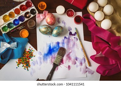 Preprimary painting of Easter Eggs, Brushes, Paints, Eggs, Sweets, Ribbons on the Dark Wooden Table. The concept of Easter. Flat lay, top view.