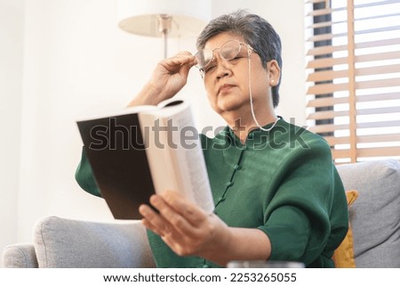 Presbyopia, Hyperopia mature asian woman holding eyeglasses having problem with vision problem trying to read text on book, eye disease of old elderly sitting on couch. Poor eyesight, health care.