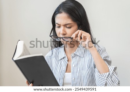 Presbyopia, hyperopia middle aged asian woman holding eyeglasses problem with vision blurred, female stare trying to read text on book at home, eye disease of old, eyesight farsightedness, health care