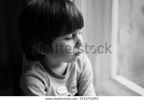 Preschool boy looking outside his home window.\
Authentic black and white portrait of a cute kid indoors. Candid\
approach with selective\
focus.
