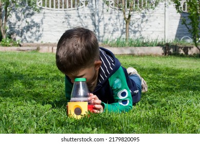 Preschool Boy Lying On His Stomach On Green Lawn Examines An Insect Using An Educational Toy Flask With Magnifying Glasses. Child Explores World Around Him With Interest. Spring. Nature.