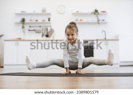Preschool cute little girl practicing yoga, standing in crane exercise, bakasana pose, working out on mat wearing sportswear, indoor full length, in white loft kitchen background.