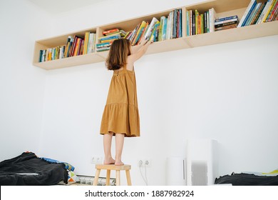 Preschool Girl Got Up On A Stool To Get A Book Off The Shelf. High Quality Photo. Real Life Moment