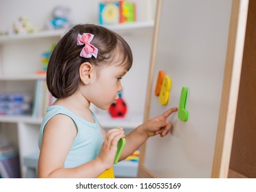 Preschool Girl Studying The Letters Of The Alphabet. Child Reaching Up At The Board With Bright Letters In A White And Bright Room. Early Development. Preparation For School.