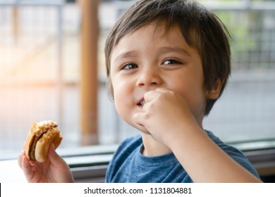 Preschool Kid Boy Eats Hamburger Sitting In Nursery Cafe,Cute Happy Boy Holding Hamburger And Looking At Camera With Smiling Face,Child Eating Delicious Homemade Burger,