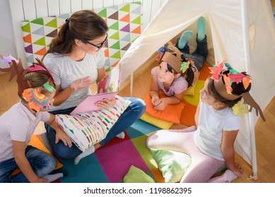 Preschool Teacher Reading A Story To Children At Kindergarten. Mother Reading To The Children.