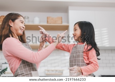Preschooler Asian kid decorating homemade cake with cream and colourful sprinkle. Delighted young mother smiling looking at daughter. Girls cooking baking spending time together in kitchen.