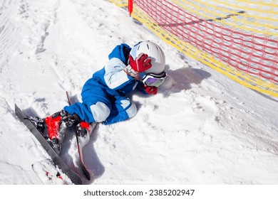 preschooler boy in a helmet, goggles, skis and winter overalls learns to ski, fell and lies on the snow. The tired child lay down on the snow to rest. Winter fun. winter holidays on a sunny cold day