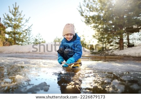 Preschooler boy is playing with a stick in brook on sunny day. Child having fun and enjoy a big puddle. All kids love play with water. Happy childhood. Outdoor activity for baby in early sping time