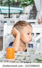 Preschooler Boy Sits In Restaurant Waiting For Order To Start Eating. Fair-haired Child Looks With Bored Expression Sitting At Wooden Table