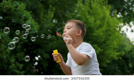 Preschooler boy spends time blowing soap bubbles creating playful atmosphere slow motion. Playing in summer city park cheerful boy happily blows bubbles. Young boy delights in blowing soap bubbles - Powered by Shutterstock