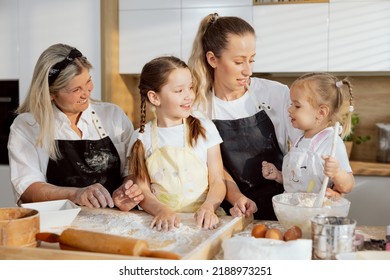 Preschooler Daughter Wearing Apronh Holding Spoon In Arm Telling Own Recipe Of Kneading Dough For Cooking Baking Cookies Homemade Pizza. Elderly Granny Mom And Older Sister Listening Smiling.