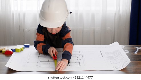 Preschooler in hardhat measures dimensions with tape measure on apartment drawing. Boy imitates role of engineer taking measurements with interest using tool on table - Shutterstock ID 2321144477