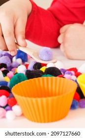 Preschooler playing with small colorful pompoms, wooden spoon and cups. Development of kids motor skills, coordination, creativity and logical thinking