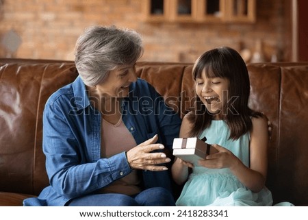 Present for beloved grandchild. Small latin girl sit on couch scream in delight receive surprise in wrapped box from smiling elderly granny. Happy hispanic grandma congratulate preteen kid on birthday