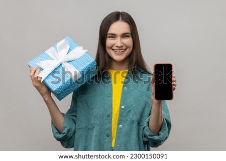 Present, bonus for mobile user. Woman holding gift box and cell phone with empty display for online shopping advertising, wearing casual style jacket. Indoor studio shot isolated on gray background.