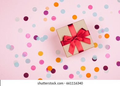 Present Box With Red Bow On Pastel Pink Background With Multicolored Confetti. Flat Lay Style.