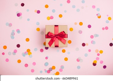 Present Box With Red Bow On Pastel Pink Background With Multicolored Confetti. Flat Lay Style.