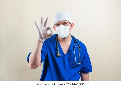 Present Doctor In Blue Suit And Protective Breathing Mask, Glasses And Stethoscope Showing Peace Sign OK By His Hand