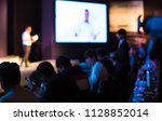 A Presenter with Hands Up Giving Presentation while Audience People Watch in Conference Hall Auditorium. Blurred De-focused Unidentifiable Presenter and Audience. Presentation Screen Video. Education.