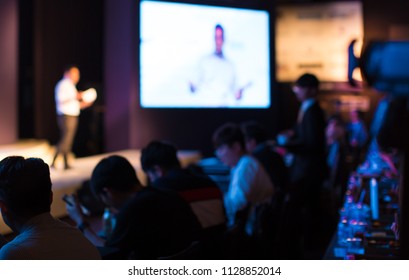 A Presenter With Hands Up Giving Presentation While Audience People Watch In Conference Hall Auditorium. Blurred De-focused Unidentifiable Presenter And Audience. Presentation Screen Video. Education.