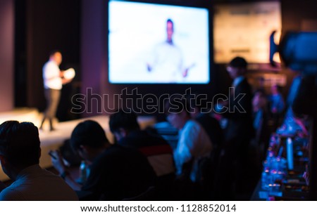 A Presenter with Hands Up Giving Presentation while Audience People Watch in Conference Hall Auditorium. Blurred De-focused Unidentifiable Presenter and Audience. Presentation Screen Video. Education.