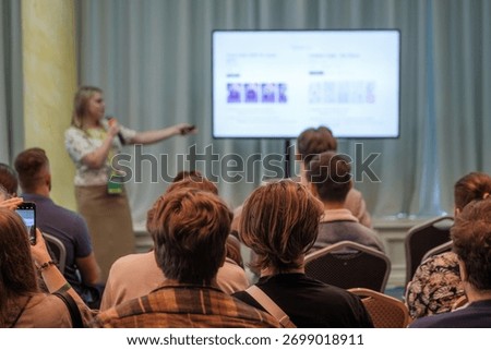 Presenter speaks into microphone while pointing to projected slide as attendees watch. Bright room, conference setting, engaged audience, learning vibe, professional presentation, sharing information.